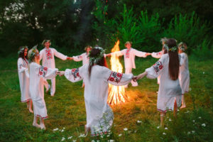 A group of young women stand in a circle holding hands around a fire, wearing white dresses with red detailing and crowns made of plants